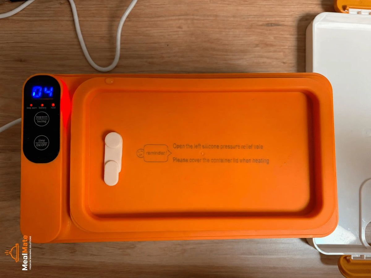 An orange silicone pressure relief valve kit by MealBot on a wooden tabletop, featuring a digital control panel with a blue LED display and a white plastic release valve.
