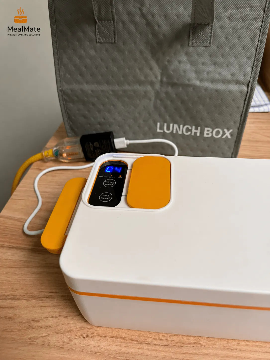 A white MealMate Lunch Box with orange accents and a digital temperature display, shown on a wooden table with its gray insulated bag and power cable in the background.